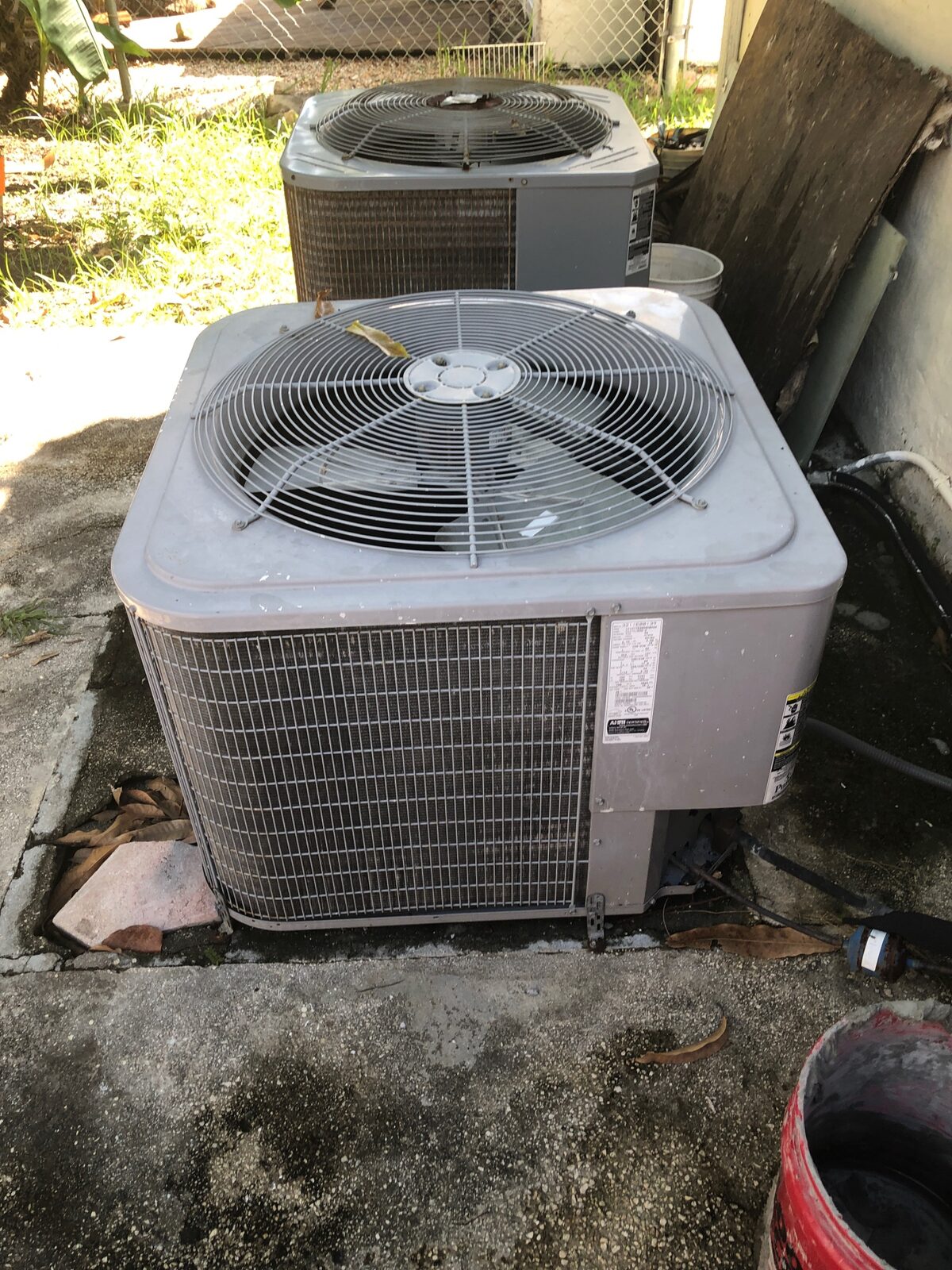 Two residential air-conditioning condensers in a leaf-strewn side yard