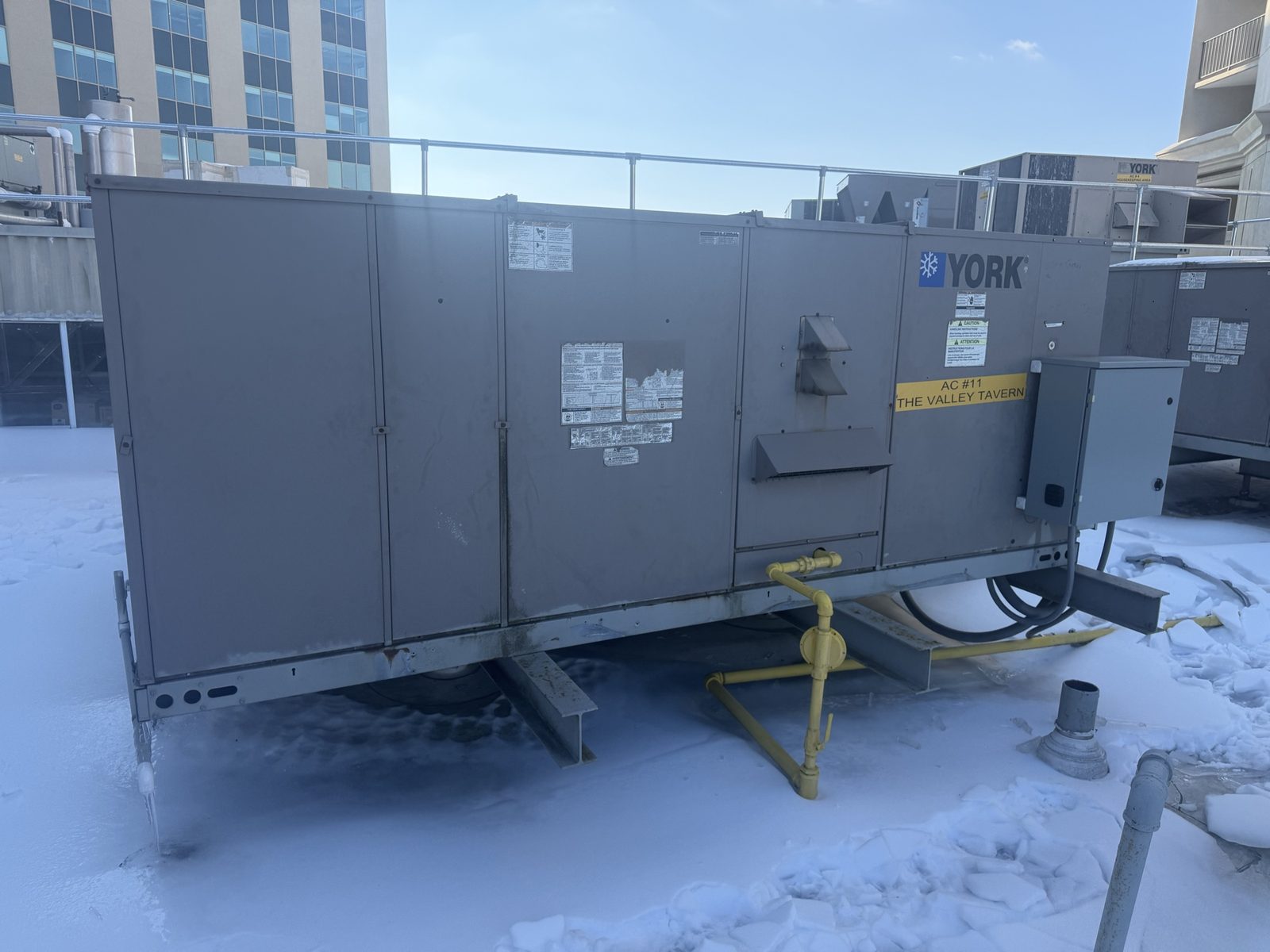A weathered gray York 20-ton commercial rooftop unit tagged 'AC #11 The Valley Tavern' sitting on steel dunnage on a snow-covered casino roof, with the casino tower behind it