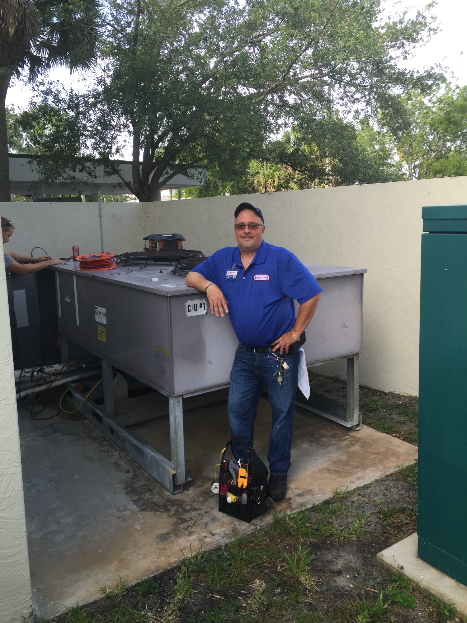 Gregory Frazier in an R. Brooks Mechanical polo and cap, standing beside a commercial condensing unit with his tool bag on the pad